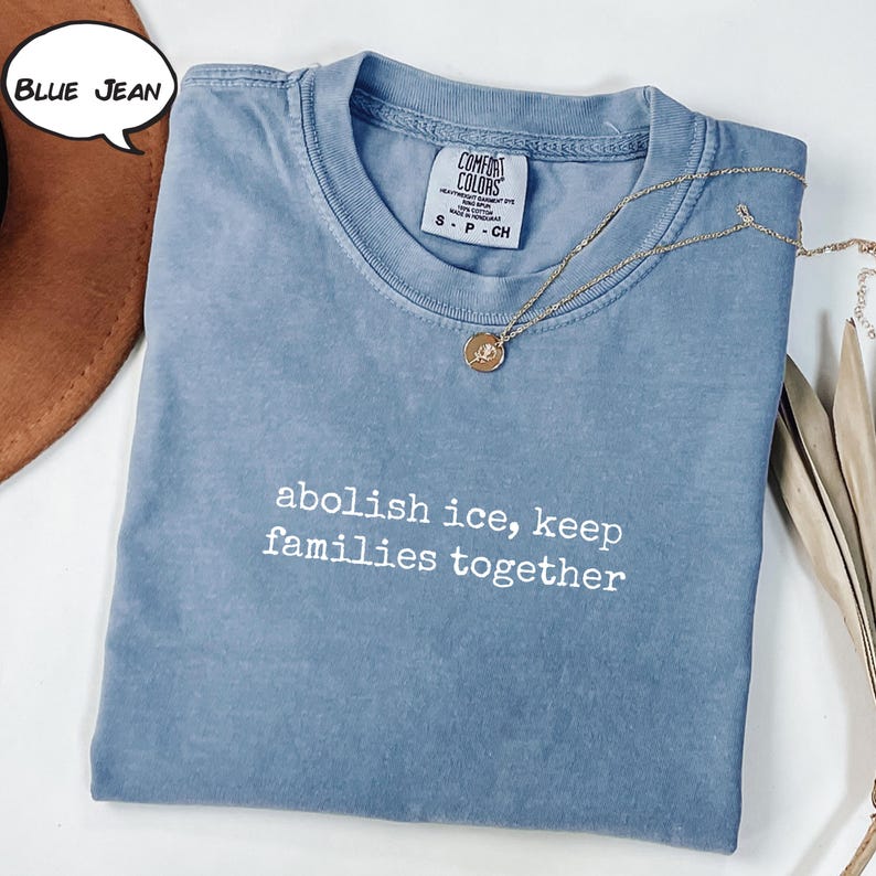 以下が含まれることがあります： 「abolish ice, keep families together」の白い文字が入った、ブルージーンズ色のTシャツ。ペンダント付きのゴールドネックレスがシャツの上にかけられています。シャツにはComfort Colorsのタグが付いています。