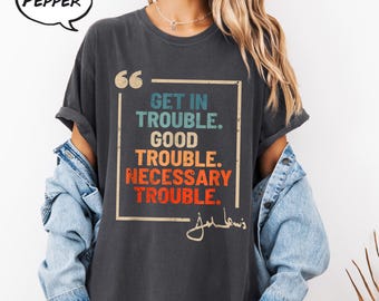 May include: Dark grey t-shirt featuring the quote "Get in Trouble. Good Trouble. Necessary Trouble." in a square frame. The text is in teal, orange, and blue. The shirt has rolled-up sleeves and is worn with a denim jacket.