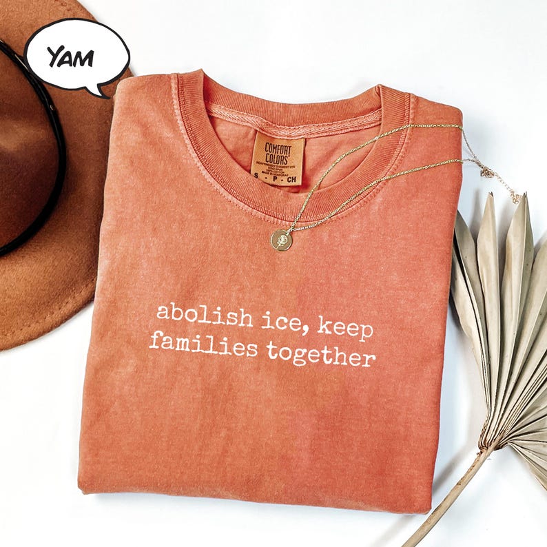 以下が含まれることがあります： 「abolish ice, keep families together」の白文字がプリントされたオレンジ色のComfort Colors Tシャツ。ペンダント付きのゴールドネックレスがシャツにかけられています。茶色の帽子と乾燥したヤシの葉が背景にあります。