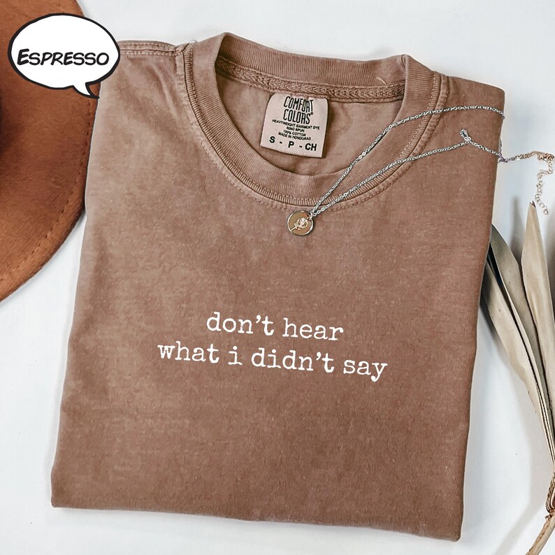 May include: Espresso-coloured t-shirt featuring the white text "don't hear what I didn't say". The shirt has a crew neck and short sleeves. A silver necklace with a small pendant is draped on the shirt. The shirt is labelled "Comfort Colors".