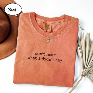 May include: An orange Comfort Colors t-shirt with the phrase "don't hear what I didn't say" in black. A gold necklace with a pendant and a brown hat are next to the shirt. A speech bubble says "YAM".