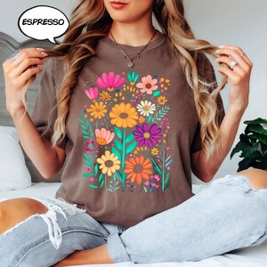 K&ouml;nnte beinhalten: Espressofarbenes T-Shirt mit einem lebendigen Blumendesign. Die Grafik zeigt bunte Blumen in Rosa, Gelb, Orange und Lila mit gr&uuml;nen Bl&auml;ttern. Das Wort "ESPRESSO" befindet sich in einer Sprechblase oben links.