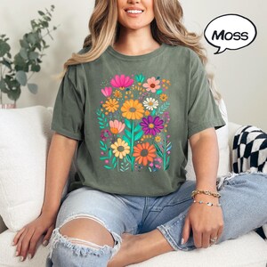 K&ouml;nnte beinhalten: Olivgr&uuml;nes T-Shirt mit einem lebendigen Blumengrafik. Das Design zeigt bunte Blumen in den Farbt&ouml;nen Rosa, Orange, Gelb und Lila. Das Wort "Moss" befindet sich in einer Sprechblase.