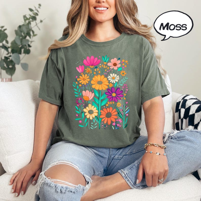 K&ouml;nnte beinhalten: Olivgr&uuml;nes T-Shirt mit einem lebendigen Blumengrafik. Das Design zeigt bunte Blumen in den Farbt&ouml;nen Rosa, Orange, Gelb und Lila. Das Wort "Moss" befindet sich in einer Sprechblase.