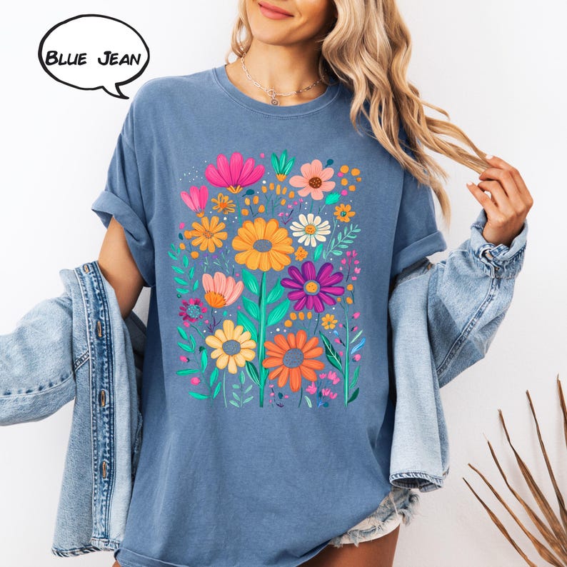 K&ouml;nnte beinhalten: Blaues Jeans-T-Shirt mit einem lebendigen Blumendesign. Die Grafik zeigt eine Vielzahl farbenfroher Blumen, darunter rosa, orange, gelbe und lila Bl&uuml;ten, mit gr&uuml;nen Bl&auml;ttern. Der Text "BLUE JEAN" befindet sich in einer Sprechblase.