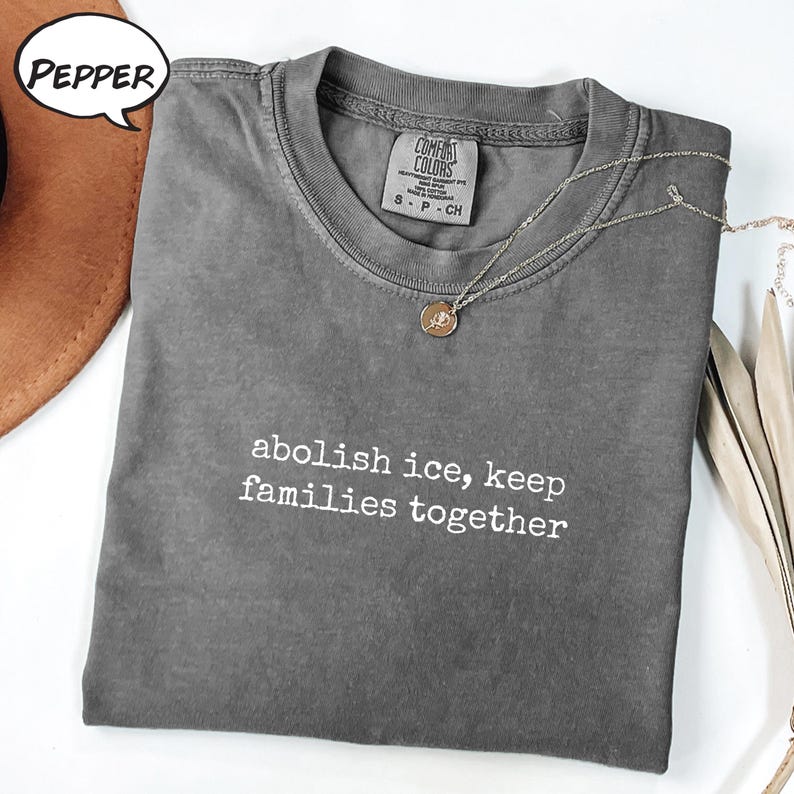 以下が含まれることがあります： 「abolish ice, keep families together」の白い文字が入ったグレーのComfort Colors Tシャツ。丸いペンダントが付いたシルバーのネックレスがシャツの上にあります。シャツは折りたたまれ、茶色の帽子と一緒に表示されています。