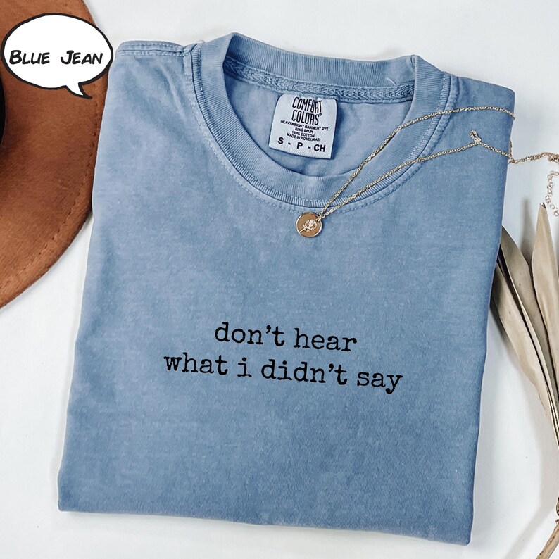 May include: A blue jean coloured t-shirt with the text "don't hear what i didn't say" in black. A gold necklace with a pendant is draped across the shirt. The shirt is folded and has a Comfort Colors tag.