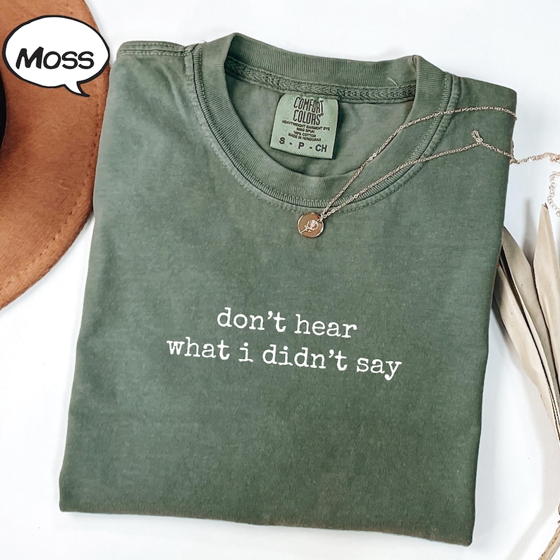 May include: A moss green t-shirt with the phrase "don't hear what I didn't say" in white lettering. A gold necklace with a pendant rests on the shirt. The shirt is folded and has a tag that says "Comfort Colors".