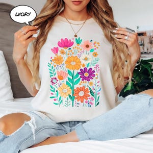 K&ouml;nnte beinhalten: Elfenbeinfarbenes T-Shirt mit einem lebendigen Blumendesign in Rosa, Orange, Gelb und Lila. Die Grafik zeigt verschiedene Blumenarten und gr&uuml;ne Bl&auml;tter. Das Shirt hat einen Rundhalsausschnitt und kurze &Auml;rmel.