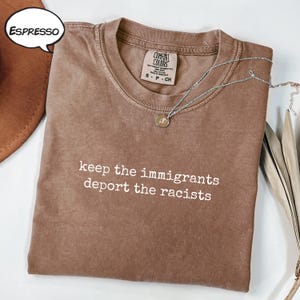 May include: Espresso-coloured t-shirt with the text "keep the immigrants deport the racists" in white. The shirt has a crew neck and short sleeves. A silver necklace with a small pendant is draped across the shirt.