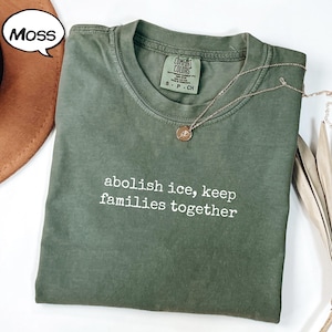 以下が含まれることがあります： モスグリーンのTシャツ。白で「abolish ice, keep families together」の文字。 金色のペンダント付きネックレスがシャツにかけられています。 シャツは折りたたまれており、Comfort Colorsのタグが付いています。