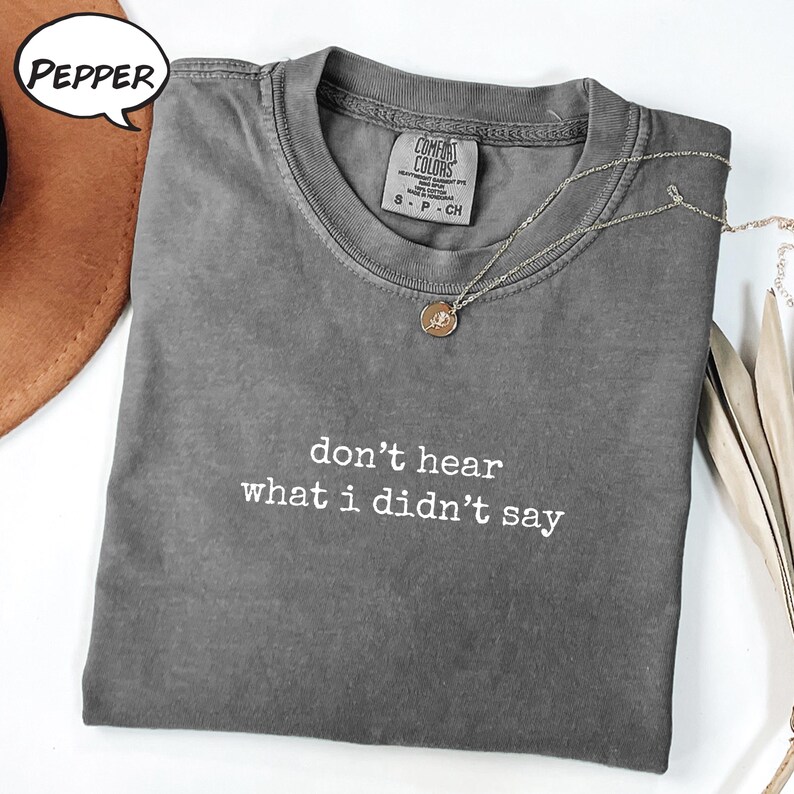 May include: A grey Comfort Colors t-shirt with the text "don't hear what I didn't say" in white. A silver necklace with a round pendant is draped across the shirt. The shirt is folded on a white surface.
