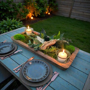 Peut inclure: Un centre de table rustique en bois rempli de mousse, de verdure et d'un morceau de bois flotté, avec deux bougies allumées dans des bougeoirs en verre. Le centre de table est posé sur une table en bois bleu, avec deux couverts.
