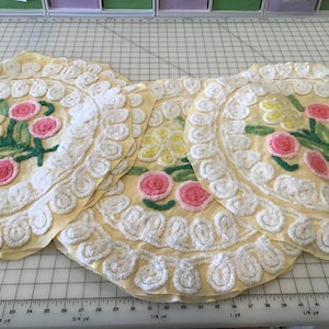 May include: Three yellow, circular placemats with embroidered floral designs. Each mat features pink and yellow flowers with green stems and leaves, surrounded by a white, textured border. The placemats are arranged on a grid surface.