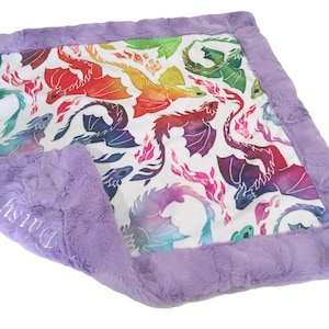 May include: A square baby blanket with a white center featuring colorful watercolor dragons. The blanket has a soft, lavender-colored plush border. The name "Daisy" is embroidered in white on the corner of the blanket.