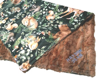 May include: A forest-themed baby blanket with a dark green top featuring woodland animals like deer, rabbits, and squirrels, and a brown faux fur underside. The blanket has the embroidered name "HAYDEN DEAN" in blue.