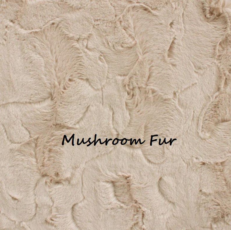 May include: Close-up of a beige faux fur fabric with a textured, wavy pattern. The words "Mushroom Fur" are printed in black at the bottom of the image. The fabric has a soft, plush appearance.