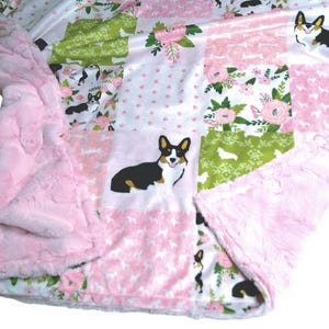 May include: A patchwork blanket featuring a repeating pattern of Corgi dogs, pink flowers, and green foliage on a white background. The blanket has a soft, textured light pink plush backing and a folded edge showing the underside fabric.