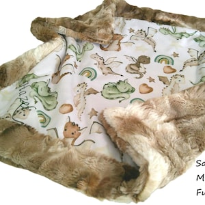 May include: A baby blanket with a white background featuring a pattern of dragons, rainbows, and stars in shades of green, brown, and tan. The blanket is trimmed with a soft, brown fur border. The text "Hazel" is visible on the blanket.