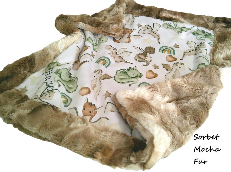 May include: A baby blanket with a white background featuring a pattern of dragons, rainbows, and stars in shades of green, brown, and tan. The blanket is trimmed with a soft, brown fur border. The text "Hazel" is visible on the blanket.