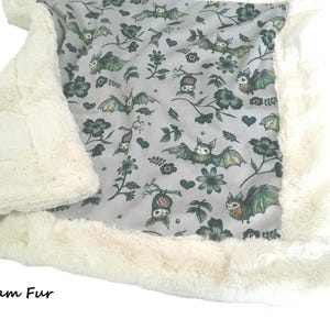 May include: A soft blanket featuring a light gray fabric with a repeating pattern of green and brown bats, dark flowers, and small hearts. The blanket is bordered by a thick, plush cream-colored faux fur trim. The text Cream Fur is visible.