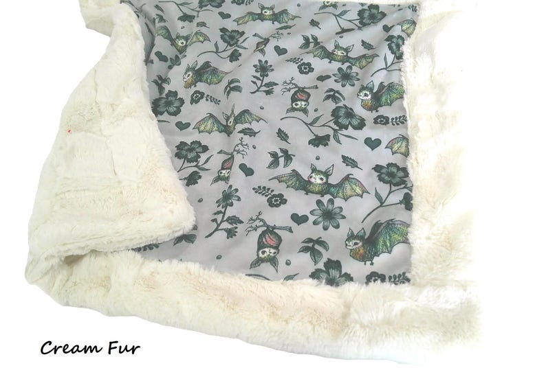May include: A soft blanket featuring a light gray fabric with a repeating pattern of green and brown bats, dark flowers, and small hearts. The blanket is bordered by a thick, plush cream-colored faux fur trim. The text Cream Fur is visible.