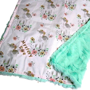 May include: A white baby blanket featuring a repeating pattern of lambs wearing flower crowns surrounded by small pink and orange flowers. The text Mystery Ewe is embroidered in brown thread. The blanket has a textured mint green plush backing.