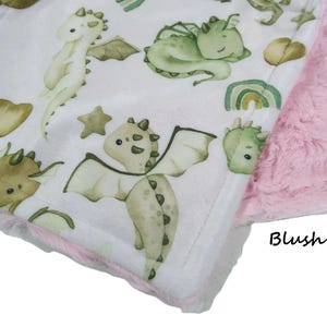 May include: A baby blanket with a white background featuring watercolor illustrations of friendly dragons in shades of green and tan. The blanket has a pink fur backing and a pink trim.