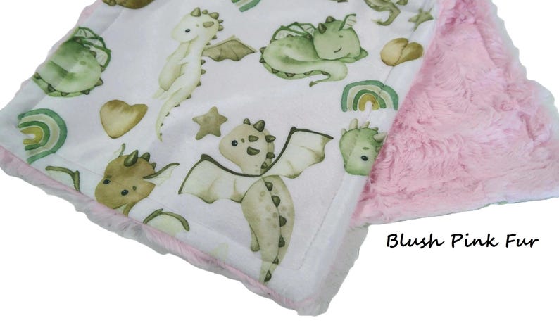 May include: A baby blanket with a white background featuring watercolor illustrations of friendly dragons in shades of green and tan. The blanket has a pink fur backing and a pink trim.