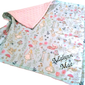 May include: A baby blanket with a light blue background featuring a repeating pattern of Alice in Wonderland characters, mushrooms, and flowers. The blanket has a soft pink dimpled minky backing and is personalized with the name Adalynn Mae.