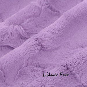 May include: Close-up of a soft, lilac-colored fur fabric. The plush material is densely textured with a wavy pattern. The words "Lilac Fur" are written in a cursive font at the bottom right corner of the image.