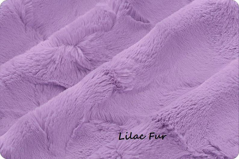 May include: Close-up of a soft, lilac-colored fur fabric. The plush material is densely textured with a wavy pattern. The words "Lilac Fur" are written in a cursive font at the bottom right corner of the image.