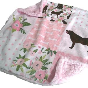 May include: A soft, plush blanket featuring a patchwork design with panels of pink and white. The pattern includes illustrations of brown dogs, pink floral arrangements with green leaves, white dog bones, and small pink paw prints on white.