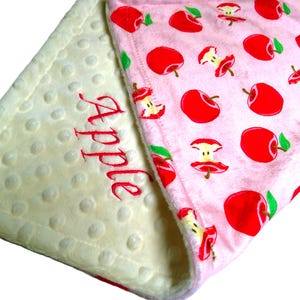May include: A white and pink baby blanket with a soft, textured side and a side with a pink background and a repeating pattern of red apples. The white side has the word "Apple" embroidered in red.
