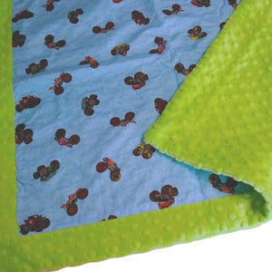 May include: A baby blanket with a light blue background featuring a pattern of monster trucks in various colors. The blanket has a bright green, textured border and backing.