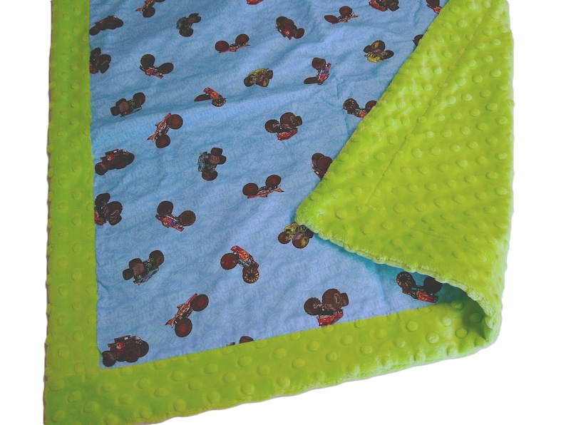 May include: A baby blanket with a light blue background featuring a pattern of monster trucks in various colors. The blanket has a bright green, textured border and backing.