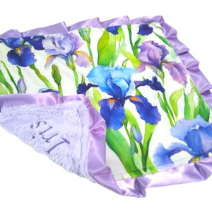 May include: A baby blanket featuring a floral iris design in shades of blue, purple, and green on a white background. The blanket has a soft, lavender plush backing and a satin border.