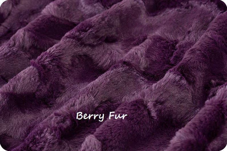 Puede incluir: Primer plano de una tela de felpa de color morado oscuro. La tela tiene una textura suave y peluda y un patr&oacute;n ligeramente acolchado. El texto "Berry Fur" est&aacute; impreso en la tela.