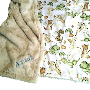 May include: A soft, beige baby blanket with a plush, textured surface. The blanket features a white patterned side with cartoon dragons in shades of green, brown, and tan. The name "Azael" is embroidered in blue on the beige side.