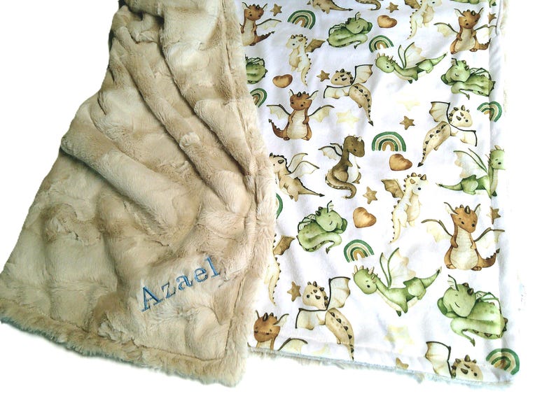 May include: A soft, beige baby blanket with a plush, textured surface. The blanket features a white patterned side with cartoon dragons in shades of green, brown, and tan. The name "Azael" is embroidered in blue on the beige side.