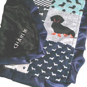 May include: A patchwork baby blanket featuring a black plush side embroidered with the name CHARLIE and a navy blue satin ruffle border. The patterned side displays various dachshund dog prints in black, white, and teal on different fabric squares.