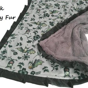 May include: A blanket featuring a light gray fabric with a repeating pattern of bats, flowers, and hearts, bordered by a dark gray ruffled satin trim. The reverse side is made of dark gray fur, as indicated by the text Dark Gray Fur.