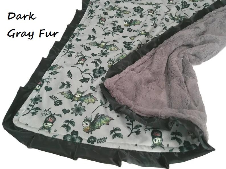 May include: A blanket featuring a light gray fabric with a repeating pattern of bats, flowers, and hearts, bordered by a dark gray ruffled satin trim. The reverse side is made of dark gray fur, as indicated by the text Dark Gray Fur.