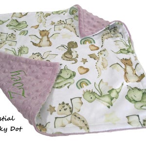 May include: A baby blanket with a white background featuring watercolor dragons in shades of green, brown, and tan. The blanket has a lavender minky dot border and the name "Zoey" embroidered in green.