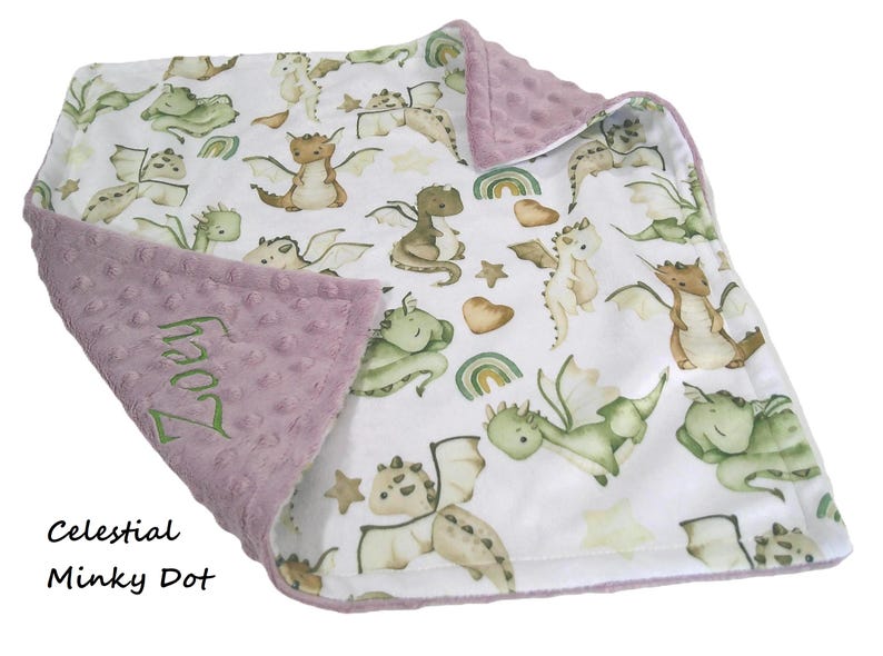 May include: A baby blanket with a white background featuring watercolor dragons in shades of green, brown, and tan. The blanket has a lavender minky dot border and the name "Zoey" embroidered in green.