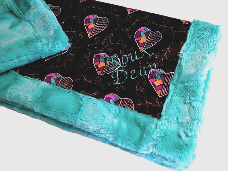 Pu&ograve; includere: Una coperta per bambini turchese e nera con un motivo a cuore e il nome "Roux Dean" ricamato in turchese. La coperta ha un bordo morbido e felpato turchese.