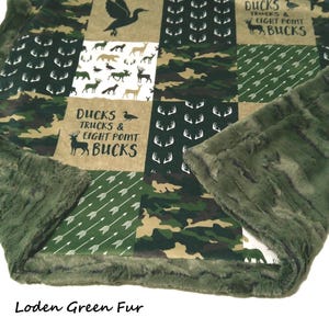 May include: A green and brown patchwork blanket with a soft, green fur trim. The blanket features squares with camouflage, deer silhouettes, arrows, and the text "DUCKS TRUCKS & EIGHT POINT BUCKS". The text "Loden Green Fur" is at the bottom.