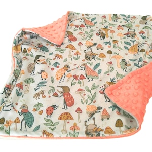 May include: A baby blanket with a whimsical forest design. The blanket features a cream-colored background with illustrations of insects, mushrooms, and foliage in various colors. The border and backing are a soft, textured coral color.