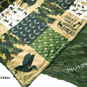May include: A patchwork blanket with a green fur backing. The top features various patterns including camouflage, arrows, and animal silhouettes. Text includes "DUCKS TRUCKS & EIGHT POINT BUCKS" and the name "HUDSON".