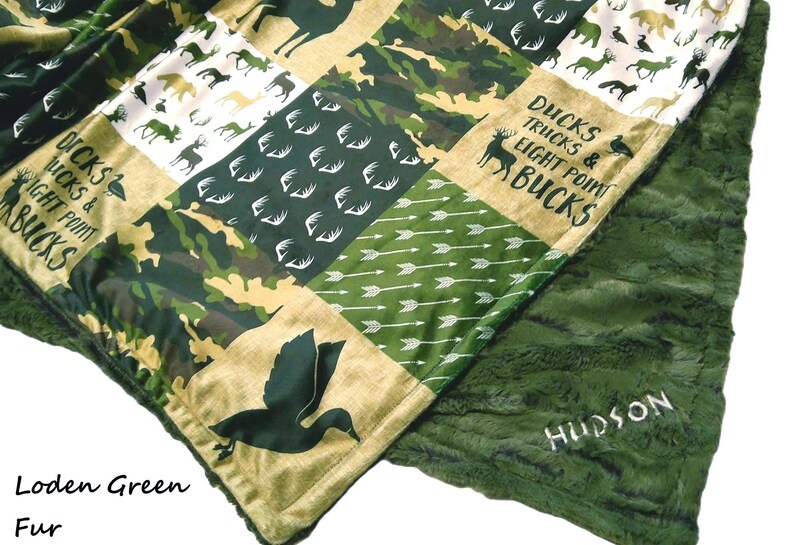 May include: A patchwork blanket with a green fur backing. The top features various patterns including camouflage, arrows, and animal silhouettes. Text includes "DUCKS TRUCKS & EIGHT POINT BUCKS" and the name "HUDSON".